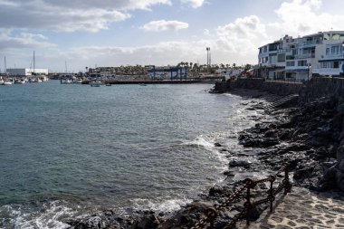 Lanzarote 'deki sahil kasabasının manzarası marinayı, tekneleri ve bulutlu bir gökyüzünün altındaki engebeli kıyı şeridini gösteriyor..