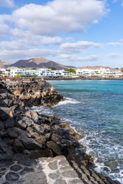 Lanzarote 'de sakin turkuaz suları ve arka planda beyaz binaları olan kayalık bir kıyı şeridi manzarası.