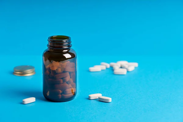 Tablets or pills of dietary supplement or vitamins in dark brown glass bottle container against blue background aimed to prevent disease. Image with copy space, horizontal. Modern color concept