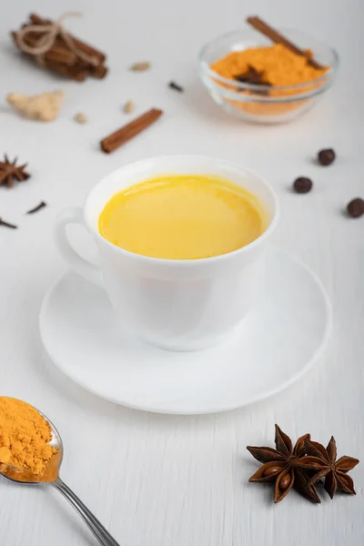 Cup full of golden or turmeric milk which is healthy, healing drink having anti-inflammatory properties served on plate with spoon of curcuma and anise on white wooden background. Vertical orientation