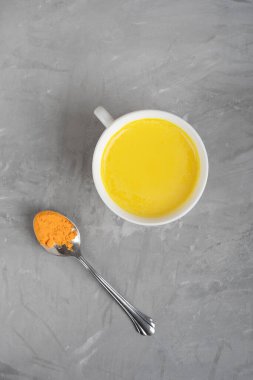 Hindistan yapımı ev yapımı bir içecek olan altın ya da zerdeçal sütü, gri beton arka planda bir kaşık curcuma tozuyla servis edilen bağışıklığı güçlendirmek için kullanılıyor. Dikey