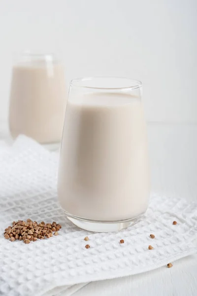 Gluten-free and lactose-free vegetarian buckwheat milk full of vitamins, minerals and antioxidants served in tall drinking glass with raw grains on textile towel on white wooden background. Vertical