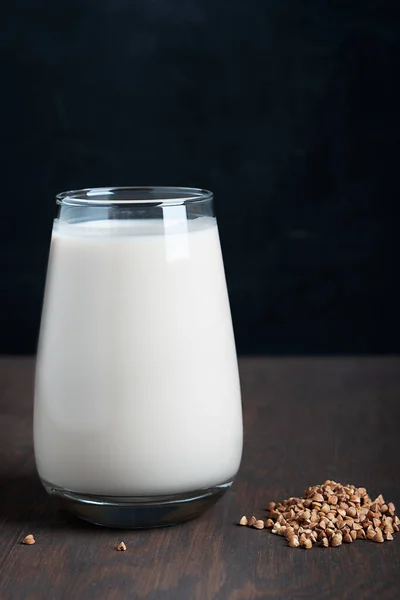 Gluten-free and lactose-free vegetarian buckwheat milk full of vitamins, minerals and antioxidants served in tall drinking glass with raw grains on dark brown wooden background at kitchen. Vertical