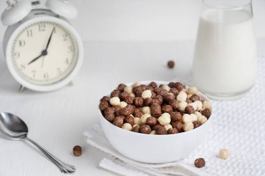 Kakao topları şeklinde hazır mısır gevrekleri ve kahvaltıya hazırlanmış bir bardak sütle dolu bir kâse dolusu beyaz ahşap masa ve sabah mutfakta çalar saat. Yatay