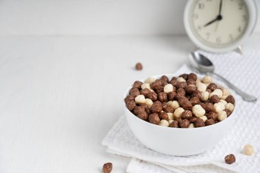 Kakao topları şeklinde hazır mısır gevreğiyle dolu bir kâse kaşık ve bir bardak sütle kahvaltı için beyaz ahşap masa üzerinde alarmlı saat ile hazırlanmış. Kopyalama alanı olan resim