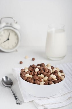Kakao ve vanilya topları şeklinde sağlıklı vejetaryen gevrekleri ile dolu bir kâse. Kahvaltı için hazırlanmış kaşık ve bir bardak sütle alarm saatiyle birlikte beyaz ahşap masada. Dikey