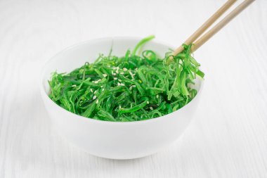 Beyaz kâsede akşam yemeği için susamlı yeşil organik chuka wakame deniz yosunu salatası. Mutfakta beyaz ahşap arka planda bambu çubuklarla yenir. Yatay yönelim resmi