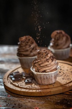 Üç tatlı ev yapımı kek ya da kahverengi kakaolu kremalı peynirle süslenmiş kek ve masanın üzerindeki tahta kesme tahtasının üzerine düşen çikolata parçacıkları. Dikey resim, seçici odak