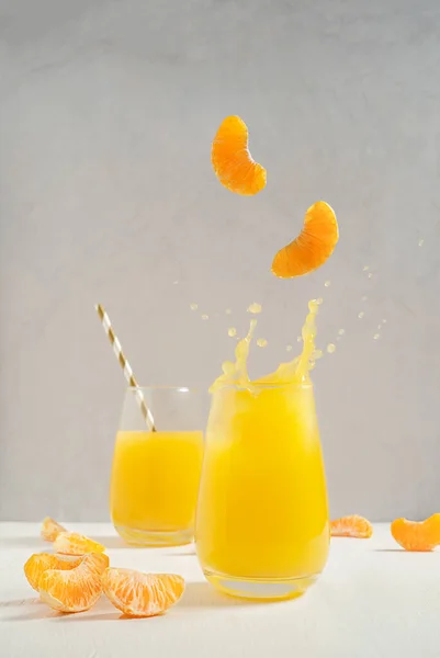 Splash of orange tangerine sweet juice served in tall drinking glass on white wooden table against gray wall at kitchen with flying of levitating sections or slices. Vertical orientation image