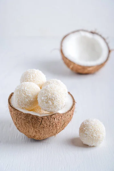 Coconut vegan sweet truffles or homemade vegetarian energy balls with a filling of condensed milk and curd cheese served in halved fresh coconut on white wooden table. Vertical image