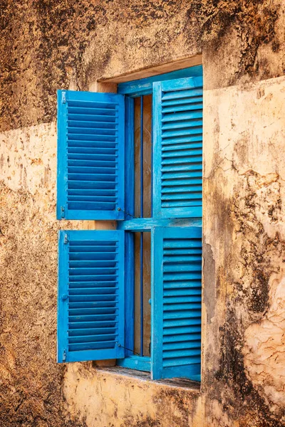 Window with opened bright blue colored weathered old fashioned shutters on yellow aged house wall of some ancient historical house. Travelling and vacation destination concept. Vertical image