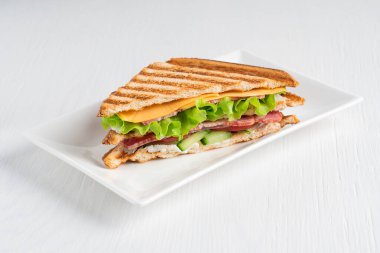 Salatalık, et ve peynirden oluşan tek bir sandviç, mutfakta beyaz ahşap arka planda servis edilen tabakta servis edilen ızgara ekmeğin arasında marul ve domates. Yatay resim