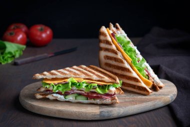 İki salatalık, bir dilim et ve peynir, marul ve domateslerden oluşan iki sandviç. Koyu kahverengi ahşap masanın üzerinde, tekstil ile servis edilen ızgara ekmek dilimleri arasında. Yatay