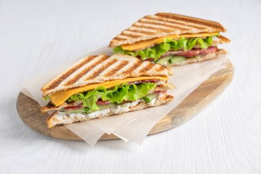 Two homemade tasty juicy sandwiches made of cucumber, meat and cheese, lettuce between slices of grilled toast bread served on cutting board on paper on white wooden table at kitchen. Horizontal clipart