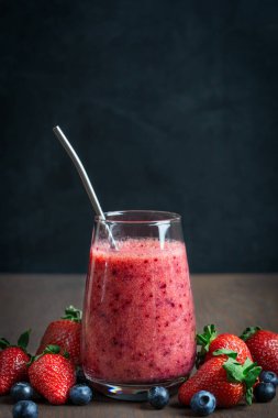 Çiğ kırmızı çilek ve yaban mersininden yapılmış taze organik smoothie ya da siyah duvara karşı koyu kahverengi ahşap masa üzerinde yeniden kullanılabilir metal pipetle içki bardağında servis edilen bataklık böğürtlenleri.