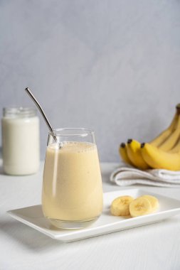 Kahvaltı için beyaz ahşap masada tabakta sıfır atık metal pipetle servis edilen içme bardağında servis edilen sütle karıştırılmış olgun muzdan yapılmış buzlu süt ya da milkshake. Dikey resim