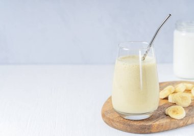 Hint Lassi içeceği ya da süt ile karıştırılmış olgun tatlı muzlardan yapılmış süt bardakta servis edilen beyaz ahşap masada metal saman ve meyve dilimleri. Kopyalama alanı olan resim
