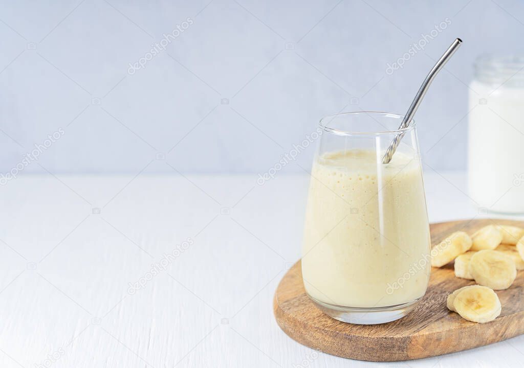 Indian lassi drink or milkshake made of ripe sweet bananas blended with milk served in drinking glass with metal straw and fruit slices on cutting board on white wooden table. Image with copy space