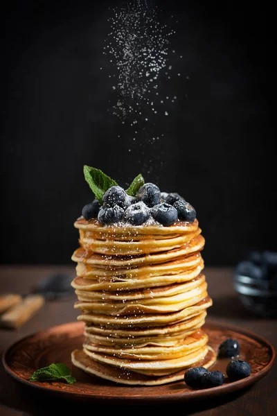 Pile of homemade organic sweet pancakes or crepes decorated with fresh blueberries and mint leaves topping served on plate against black wall on kitchen table with sugar powder sprinkles. Vertical