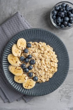 Yulaf ezmesinin en üst görüntüsü yulaf lapası, taze yaban mersiniyle süslenmiş yulaf lapası ve koyu gri tabakta dilimlenmiş muz. Sağlıklı kahvaltı için beton arka planda tekstil ile servis ediliyor. Dikey