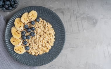 Haşlanmış yulaf ezmesinden yapılmış yulaf lapası, taze yaban mersini ve dilimlenmiş muzla süslenmiş ve kahvaltıda beton zeminde mavi gri tabakta servis edilen yulaf lapası manzarası. Kopyalama alanı olan resim