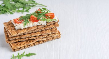 Bir yığın çıtır ekmek, içinde ricotta soslu çavdar unu, vişneli domates dilimleri ve roket yaprağı ya da beyaz ahşap zemin üzerinde roka olan düz ve kuru bir kraker. Kopyalama alanı olan resim
