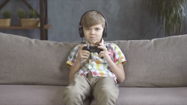 Garçon enfant paresseux jouant à un jeu vidéo avec joystick et écouteurs assis sur un canapé — Video