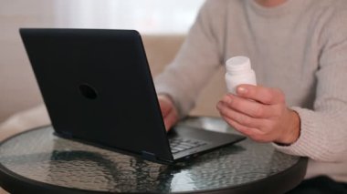 Delikanlı elleri ilaç şişesini tutuyor ve dizüstü bilgisayarına bir şeyler yazıyor..