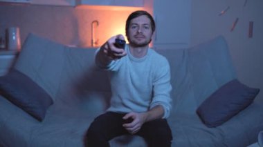 Genç adam uzaktan kumandayı tutuyor ve şaşırmış görünüyor. Evdeki kanepede televizyon izlerken.