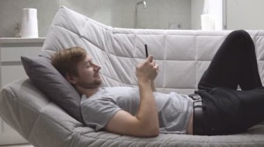 Şaşkın genç adam akıllı telefondaki cep telefonu uygulamasını kullanarak koltukta uzanıyor.