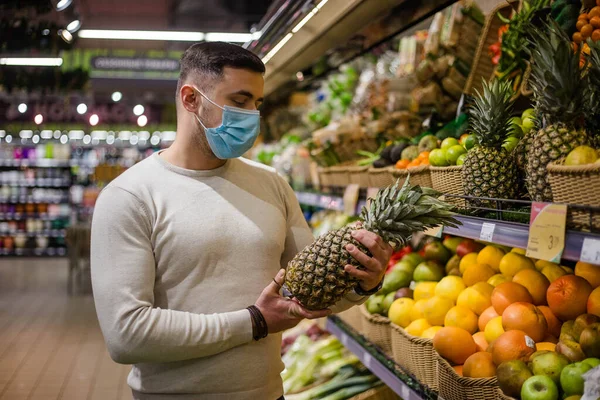 Молодой человек в маске выбирает ананас в продуктовом магазине — стоковое фото