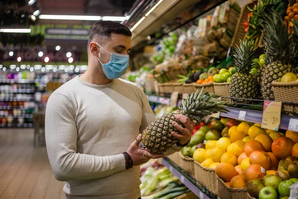 Young man in mask choosing pineapple at grocery store Stock Image