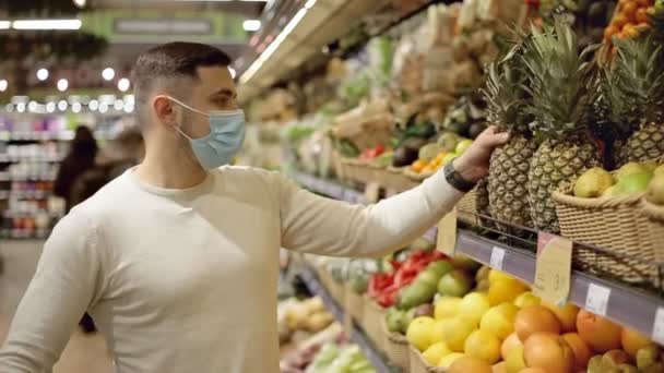 Hombre joven con máscara eligiendo piña en la tienda de comestibles — Vídeo de stock