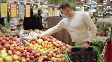 Maskeli adam yemek rafından taze bir elma alıyor. Alışveriş yapan kişi markette elma seçiyor.