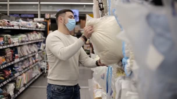 Tek kullanımlık tıbbi maske takan genç adam süpermarkette bir yastık seçiyor. Mobilya mağazasında erkek yatak alışverişi.. — Stok video