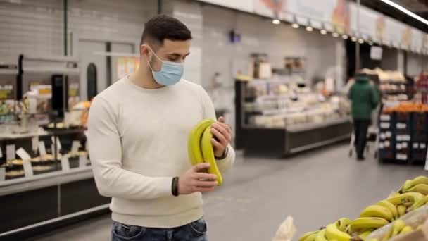 Молодой человек в маске выбирает бананы в продуктовом магазине — стоковое видео