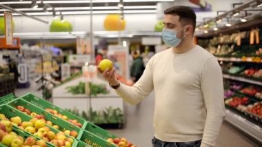 Maskeli adam yemek rafından taze bir elma alıyor. Alışveriş yapan kişi markette elma seçiyor.