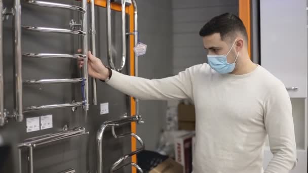 Hombre joven con máscara médica desechable elige una toalla calentada rieles en una ferretería. — Vídeos de Stock