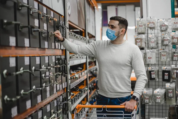 Az eldobható orvosi maszkot viselő fiatalember portréja, amint új ajtókilincseket választ a háztartási boltban. Stock Fotó