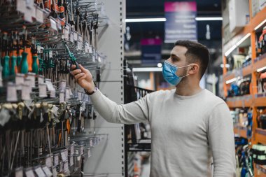 Tek kullanımlık tıbbi maske takan genç adam alet edevat dükkanından bir tornavida seçiyor.
