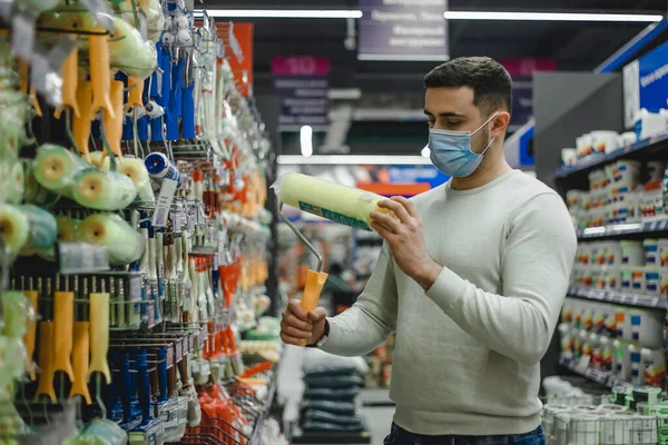Покупатель в медицинской маске выбирает ролик краски в хозяйственном магазине.. — стоковое фото