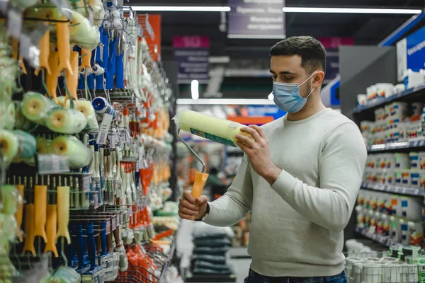 Покупатель в медицинской маске выбирает ролик краски в хозяйственном магазине.. Лицензионные Стоковые Фото