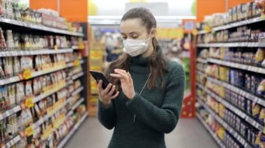 Tıbbi maske takan bir kadın markette alışveriş yaparken akıllı telefon kullanıyor.