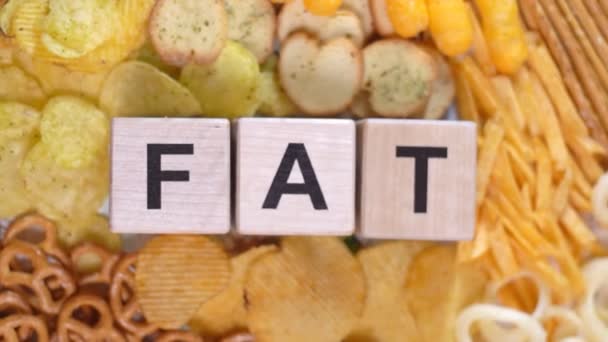 The word FAT is made of wooden cubes on a background of different unhealthy salty snacks: chips, crackers, pretzels, onion rings. Top view. Background — Stock Video