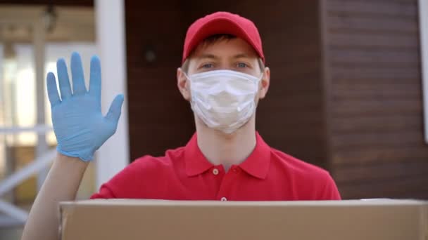 Portrait du livreur portant un masque de sécurité tenant une boîte et agitant la main. Service de livraison en quarantaine coronavirus covid-19. — Video