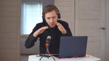 Kulaklıklı genç hippi ev stüdyosunda dizüstü bilgisayarla podcast kaydediyor, mikrofon kullanarak röportaj yapıyor. Yavaş çekim