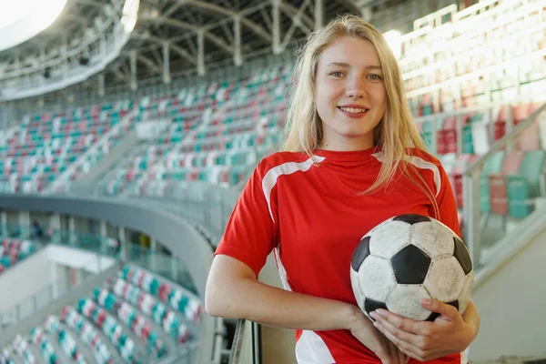 Portrét mladé bělošky v červeném fotbalovém pruhu držící míč na velkém fotbalovém stadionu, usmívající se do kamery — Stock fotografie