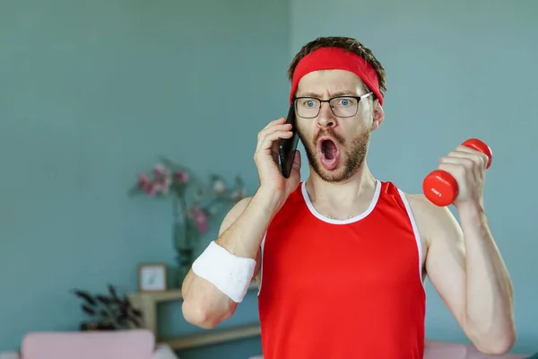 Retrato de divertido friki sorprendido hablando por teléfono mientras se ejercita con la mancuerna —  Fotos de Stock