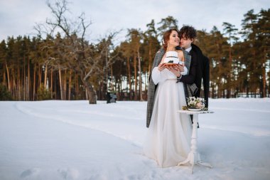 Gelin ve damat ellerinde bir düğün pastası tutuyorlar, ormanda karın içinde duruyorlar..
