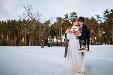 Gelin ve damat ellerinde bir düğün pastası tutuyorlar, ormanda karın içinde duruyorlar..